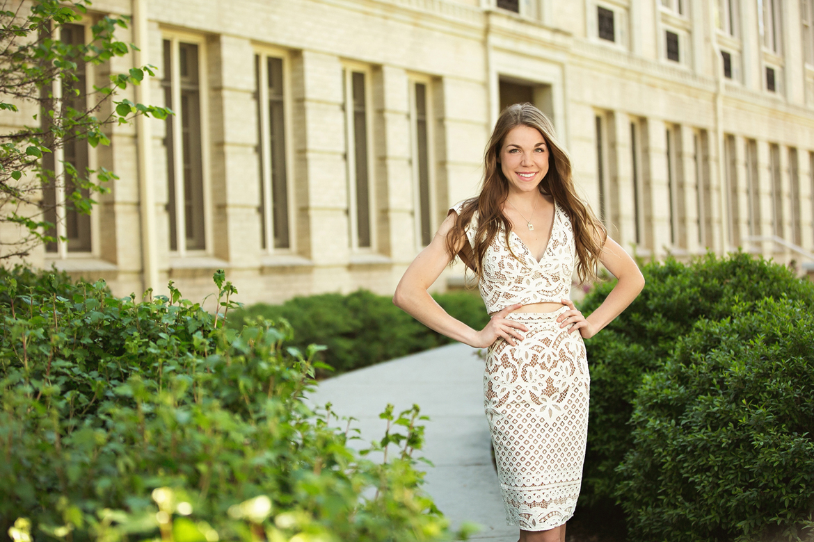 beautiful college grad standing in front of building smiling in white lace dress
