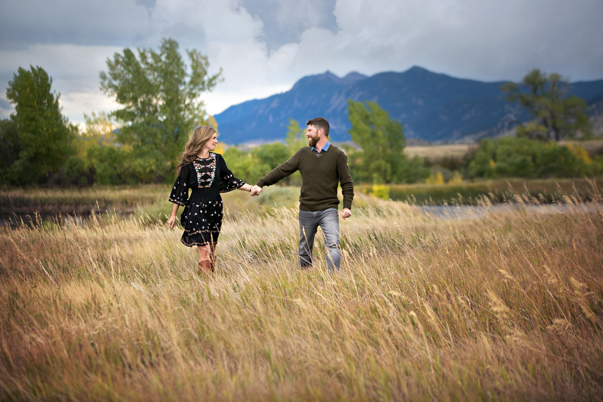 Portrait of a couple holding hands and walking while looking at each other and smiling with a scene of the flatirons in Boulder, Colorado behind them