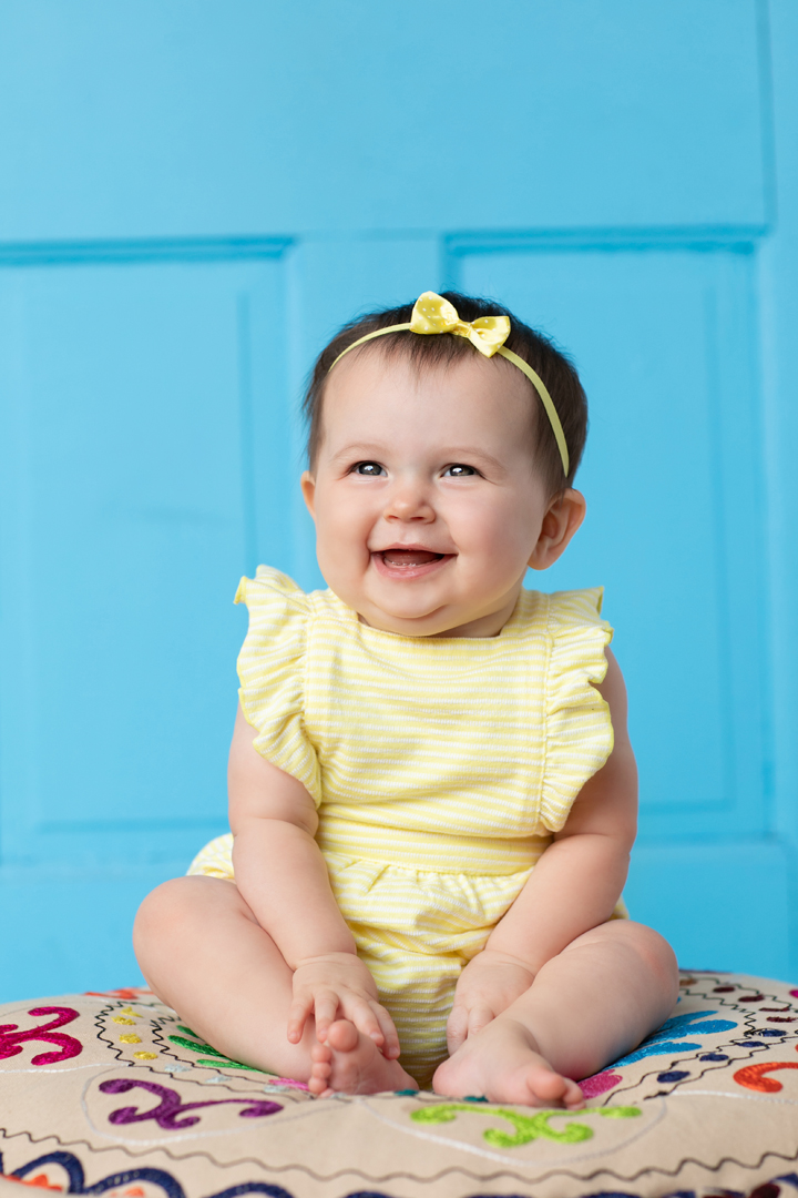 Studio portrait of a baby about 6 months old in front of a bright blue background wearing a yellow romper