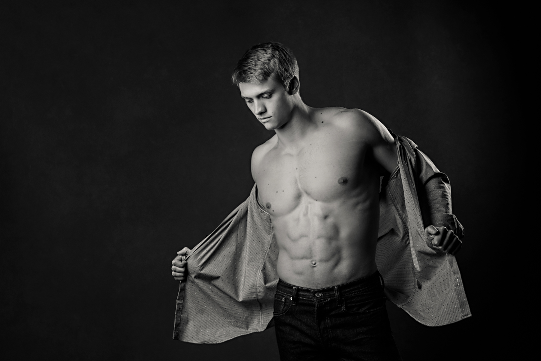 Black and white studio portrait taken of an incredibly ripped young man pulling his shirt off to reveal his chiseled abs