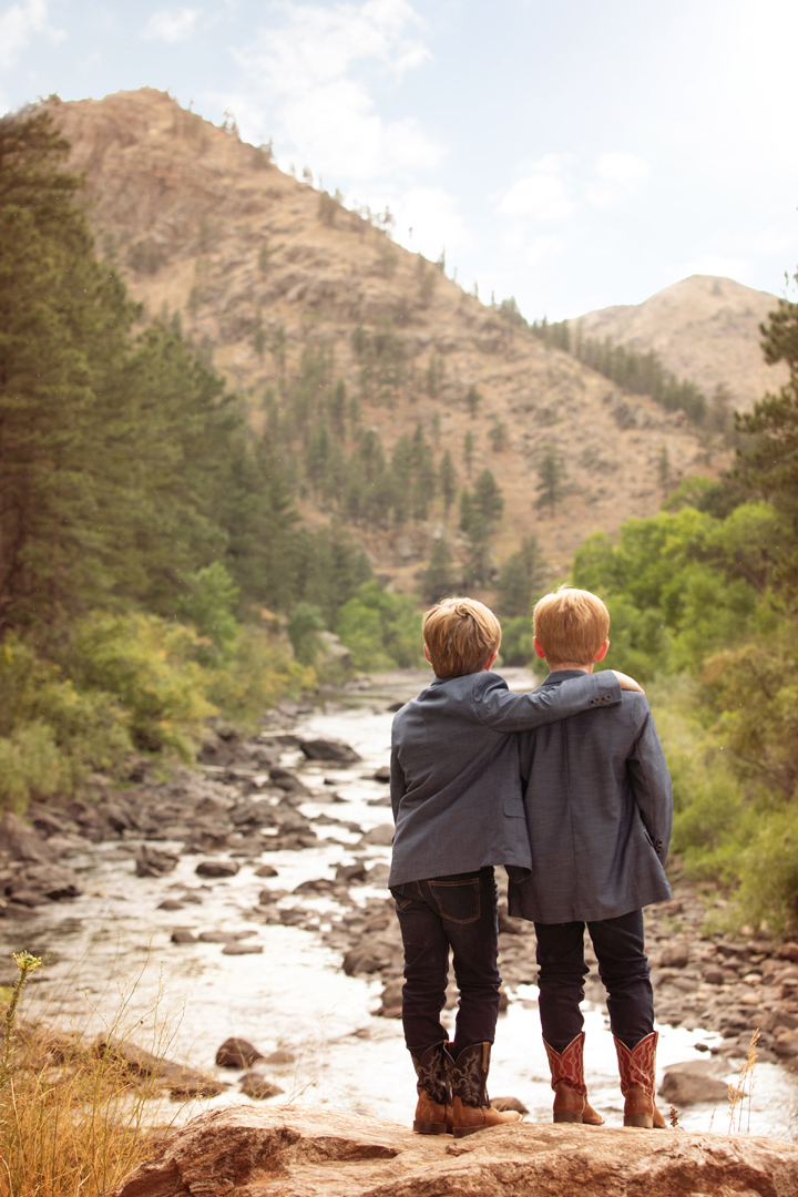 Portrait of 2 brothers wearing cowboy boots, taken from behind them with one brother wrapping his arm around his brothers' shoulder, looking at the mountain scene in the Poudre Canyon