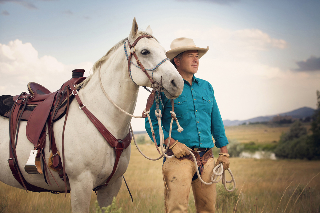 Branding photo of a saddle maker standing in field with his horse and custom saddle
