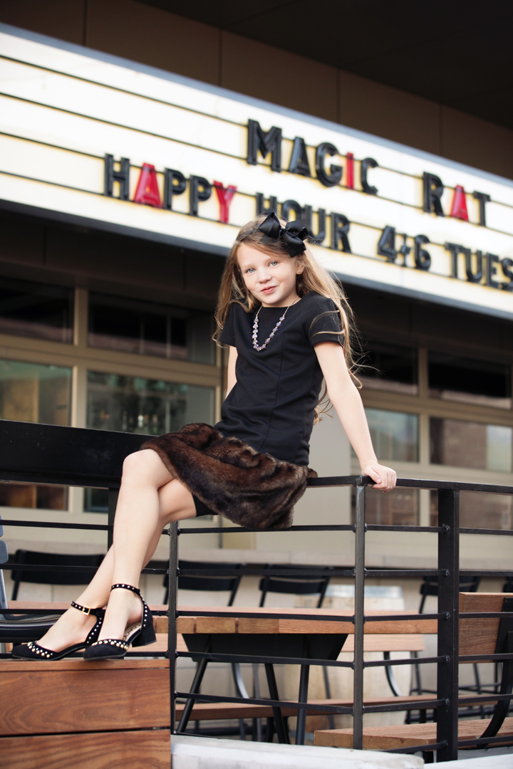 Little girl with fur skirt on and a black bow in her hair, sitting on the railing in front of the Magic Rat in Ft. Collins, CO