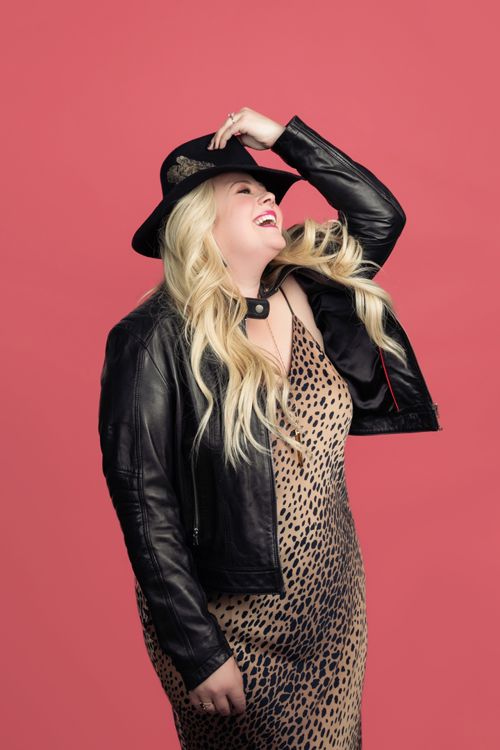 Branding photo of a personal stylist holding a fedora,  laughing on a pink background.