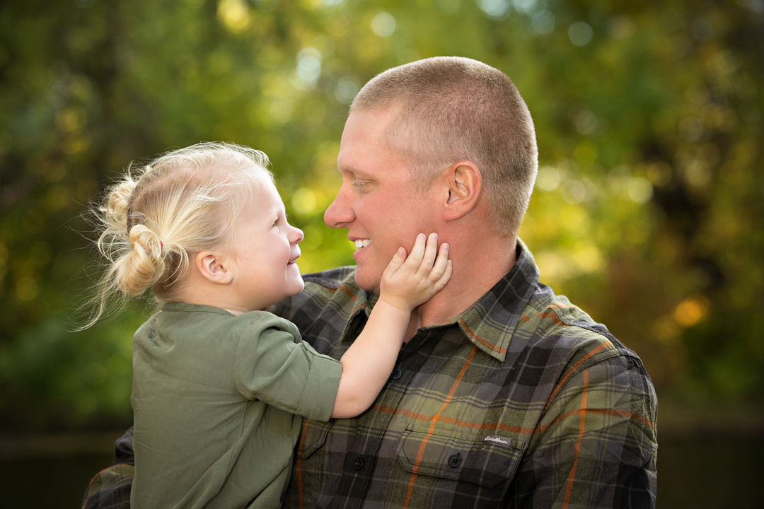 Cutest little blonde girl being held by her dad with her hand on his face, they are smiling at each other.  The background is fall colors at Legacy Park in Ft. Collins