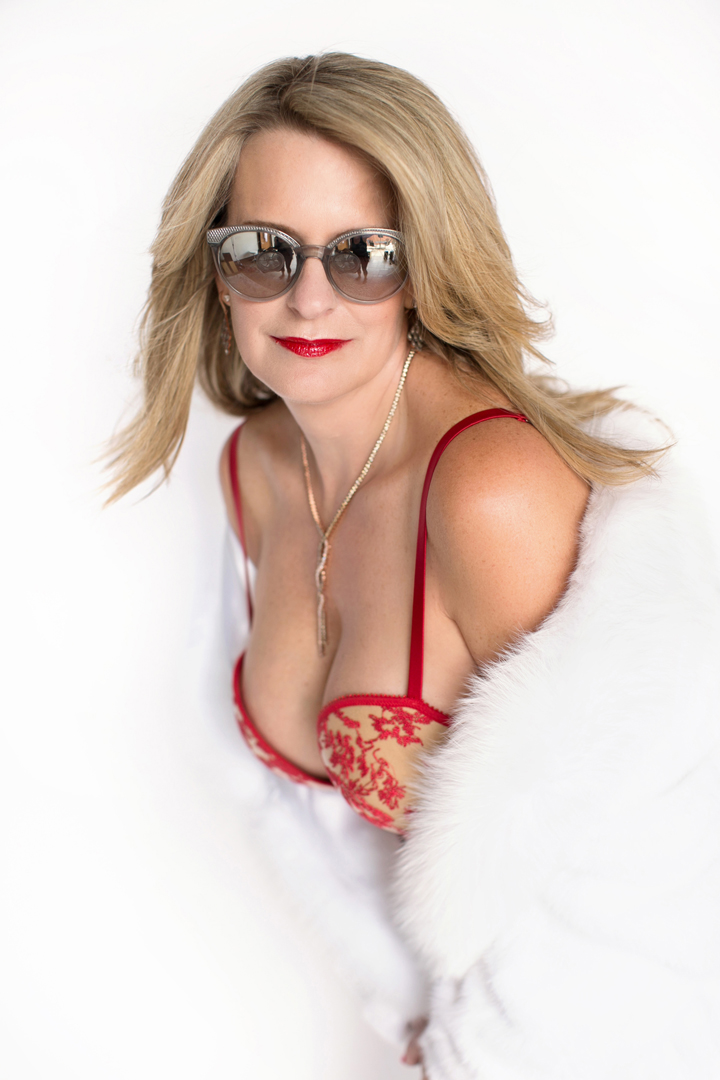 woman in red lingerie and white fur coat and sunglasses softly smiling at camera
