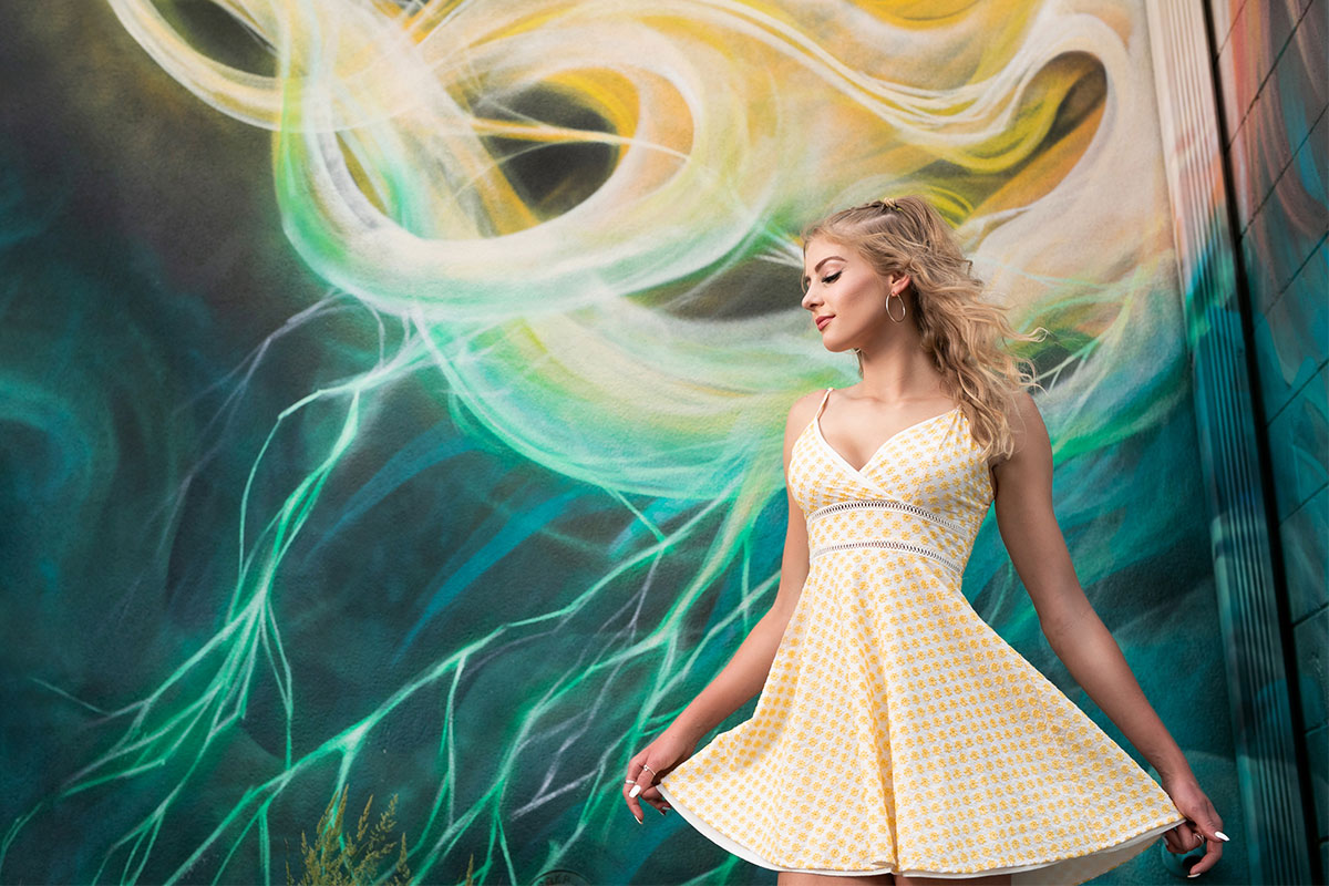 A beautiful high school senior girl poses in a yellow dress in front of a wall mural in Down Town Fort Collins, Colorado.