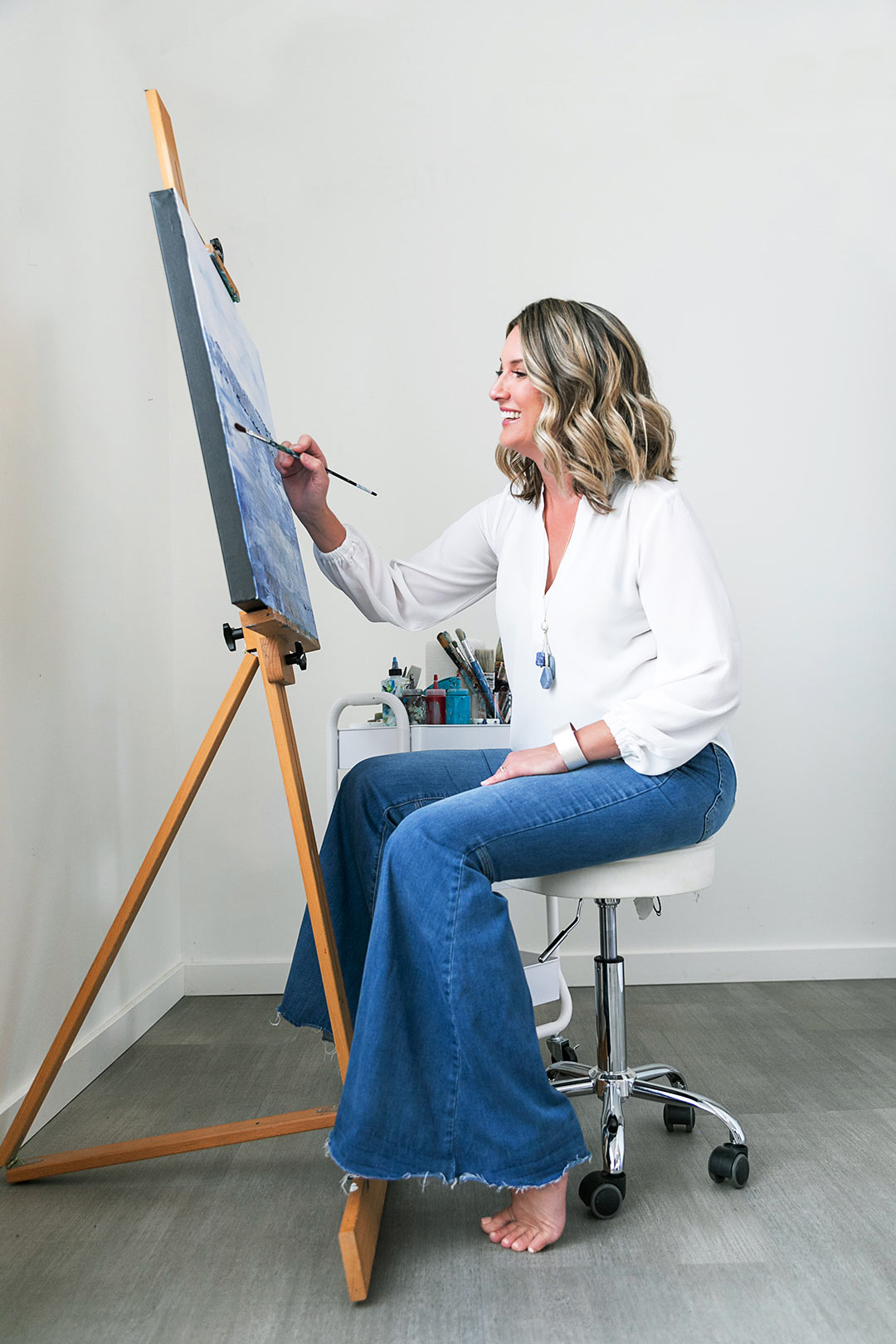 A painter smiles as she paints siting at her easel while having a lifestyle business portrait session done in a studio with Photography by Desirée