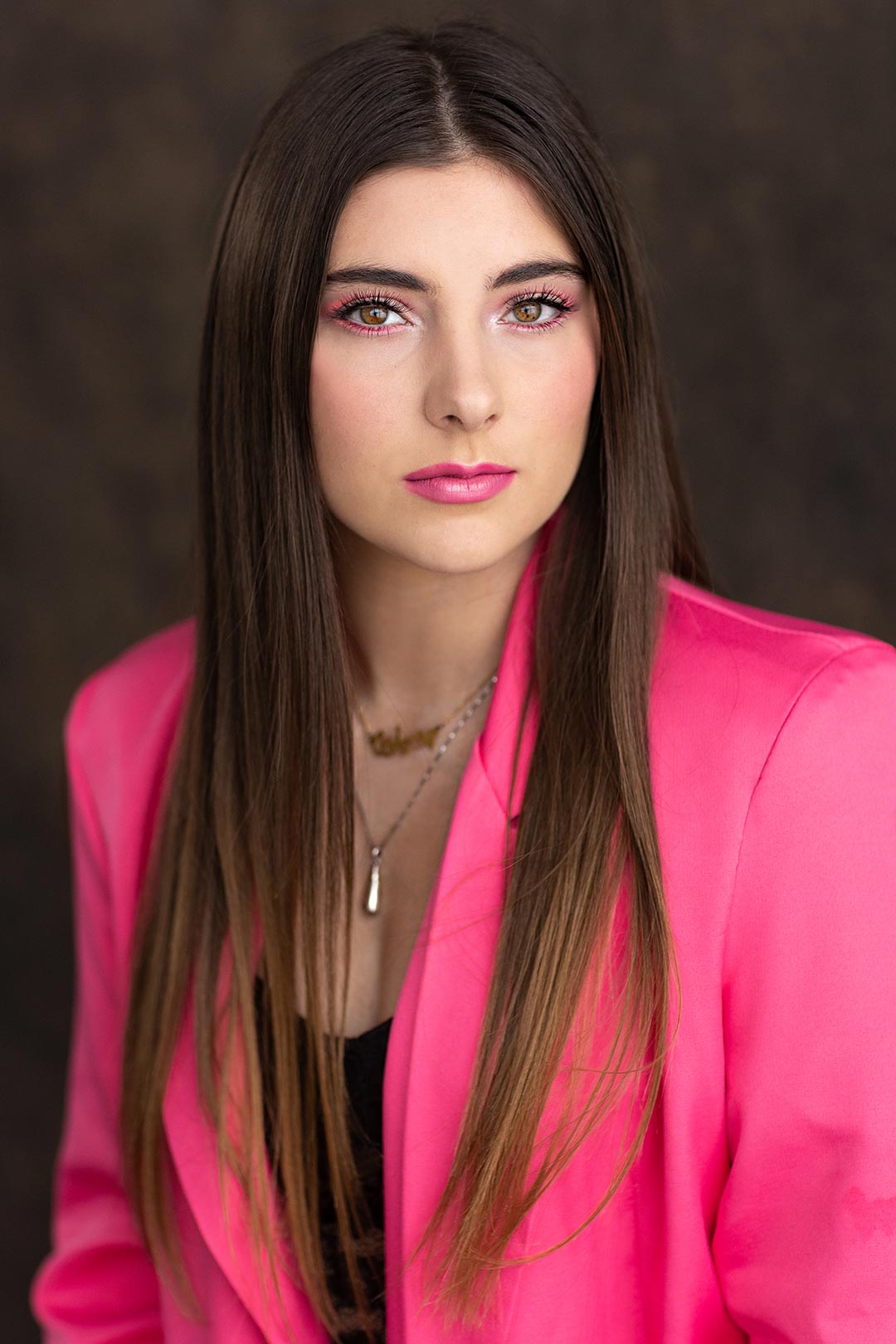 A senior girl smolders a smile in a hot pink jacket with matching lipstick and eye makeup done as a part of a themed shoot for Photography by Desirée's models team in Fort Collins Colorado. 