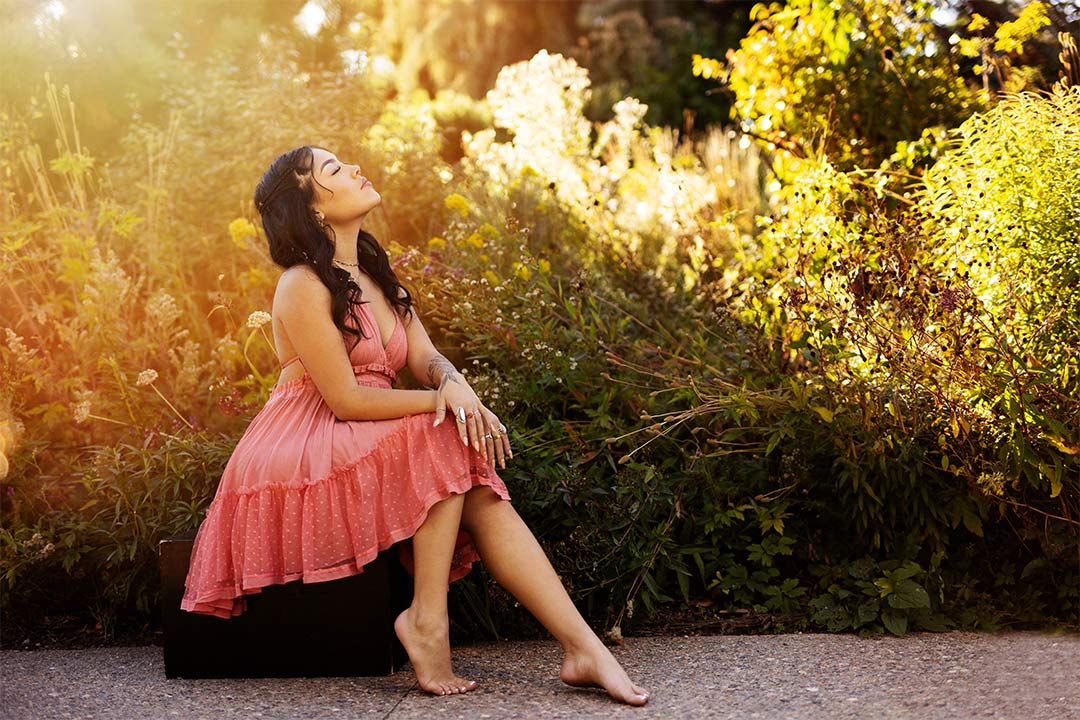Young high school senior model posing in a seated position with her face towards the sun wearing a coral colored dress in the Botanical Gardens.